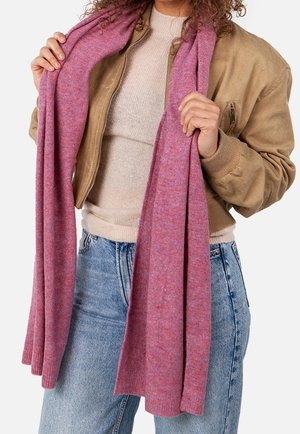 Personne tenant une longue écharpe rose, portant un pull beige, une veste marron clair et un jean bleu clair sur un fond blanc.