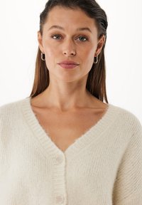 Krämfärgad cardigan med V-ringning, ribbade kanter och två stora knappar, tillverkad av mjukt, texturerat tyg med en fuzzy yta.