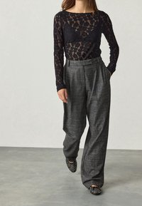 Haut à manches longues en dentelle noire sur un pantalon gris taille haute plissé avec un motif texturé. Les chaussures sont en matériau noir avec des accents cloutés.