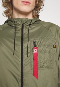 Olive green windbreaker with a hood, zippered front, chest pocket, and red fabric tag. Features black drawstrings and textured fabric.
