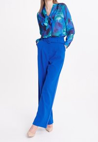 Blouse à motifs bleus avec un nœud au col, associée à un pantalon large taille haute bleu et des talons beiges. Texture lisse.