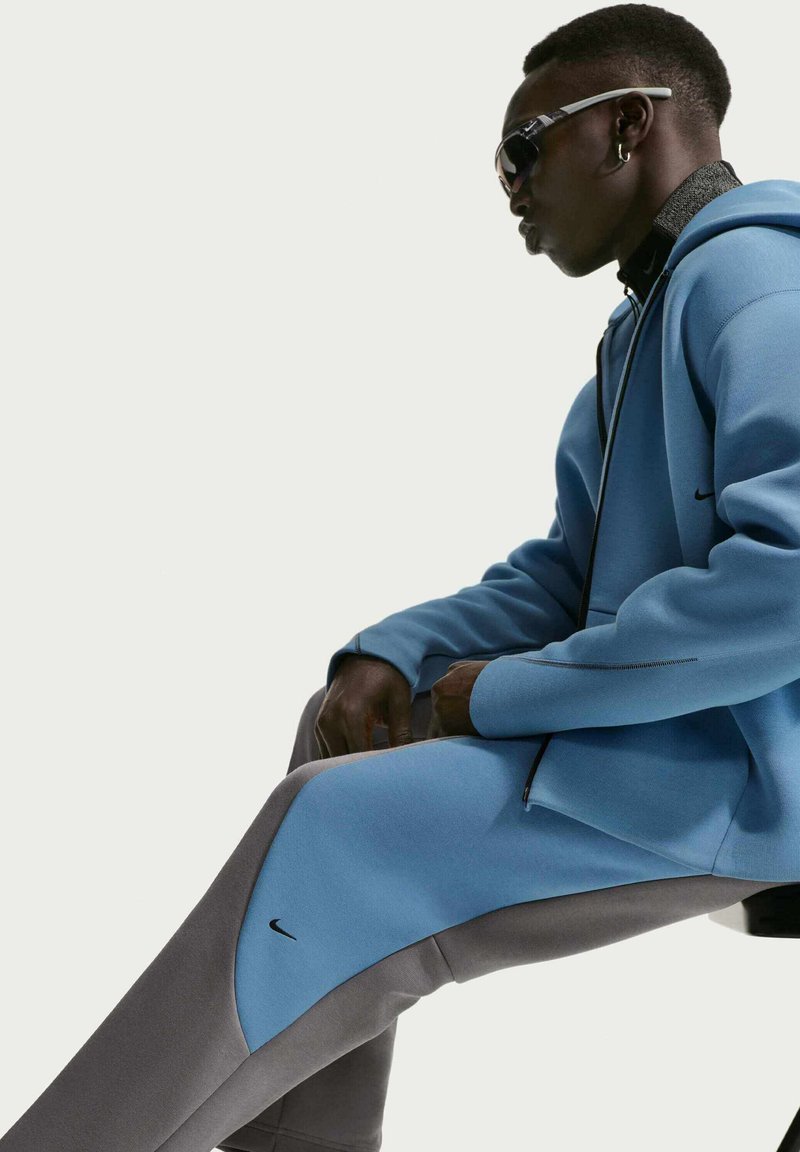 Mann in blauem und grauem Nike-Trainingsanzug und Sonnenbrille, seitlich sitzend vor hellem Hintergrund.