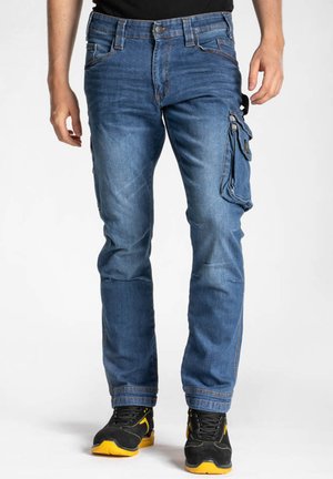 Pantalons cargo en denim bleu avec plusieurs poches, un design fuselé et des coutures visibles. Portés avec des chaussures noires et jaunes.