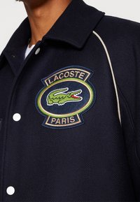 Námořnická vlněná bunda s bílými patentními knoflíky, strukturovaným povrchem a vyšitým logem Lacoste s zelenými a červenými akcenty na hrudi.