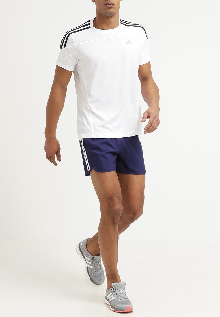 Pánský sportovní outfit se skládá z bílé košile s krátkým rukávem, námořnických šortek a šedých běžeckých bot s růžovými akcenty. Viditelné logo Adidas.