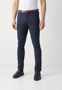 Pantalones ajustados azul marino de algodón, con un cinturón de cuero marrón y mocasines negros con detalles en dorado.
