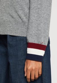 Maglione grigio lavorato a maglia con polsini a coste caratterizzati da righe rosse, bianche e blu navy. Abbinato a denim scuro, mostrando un tessuto strutturato e un design aderente.