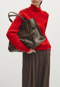 Brown leather tote bag with textured finish, held by a person in a red ribbed sweater and brown pleated trousers.