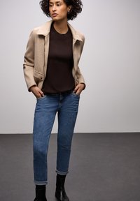 Giacca beige in misto lana con colletto a risvolto, maglione marrone e jeans blu risvoltati, abbinati a stivali neri alla caviglia. Outfit semplice e casual.