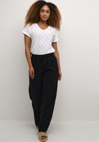 Witte V-hals T-shirt met gaatjespatroon, gecombineerd met zwarte broek met wijde pijpen, voorzien van een elastische tailleband en trekkoord. Draagt lichtgewicht sandalen.
