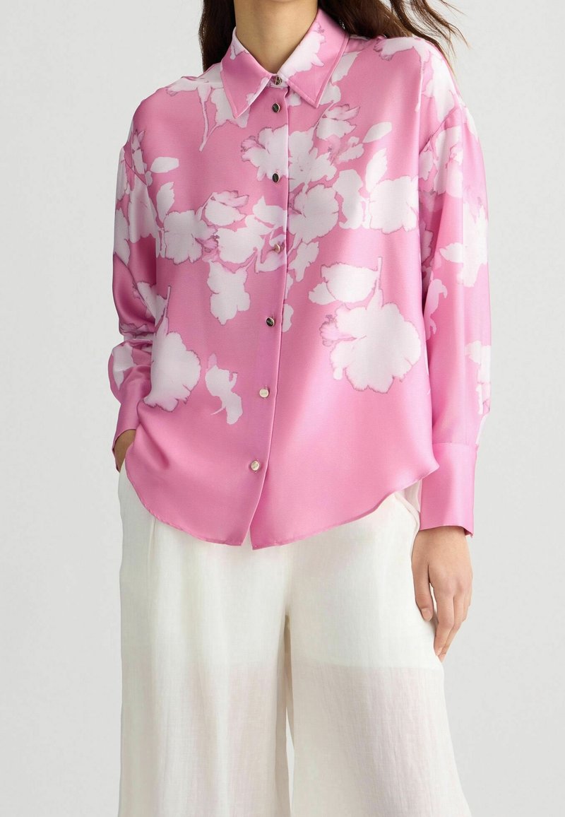 Femme portant un chemisier rose ample à boutons avec imprimé floral blanc et un pantalon blanc à jambes larges, les mains dans les poches, debout devant un fond uni.