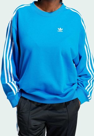 Osoba ubrana w niebieską bluzę Adidas z białymi paskami na rękawach i czarne spodnie, ręce w kieszeniach, na jasnym tle.