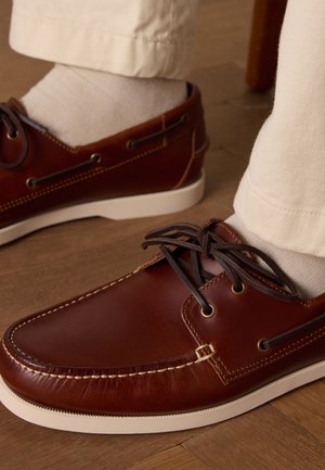 Scarpe da barca in pelle marrone con suole in gomma bianca, caratterizzate da cuciture decorative, occhielli metallici e lacci marroni. Indossate con pantaloni di colore chiaro.