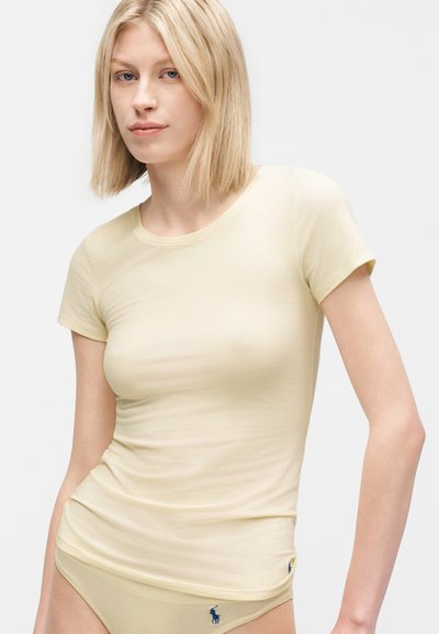 Ξανθιά γυναίκα που φοράει εφαρμοστό απαλά κίτρινο κοντομάνικο πουκάμισο και ταιριαστό εσώρουχο με μπλε λογότυπο πόλο, στέκεται μπροστά σε απλό φόντο.