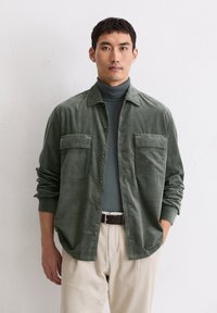 Camicia in velluto a coste verde con due tasche sul petto, chiusura con bottoni, indossata sopra un dolcevita grigio. Modello la abbina a pantaloni color crema.