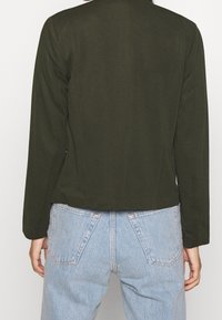 Person som bär en olivgrön långärmad topp och ljusblå högmidjade jeans, vänd bort mot en enkel vit bakgrund.