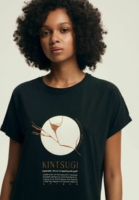 Czarny bawełniany T-shirt z okrągłym nadrukiem z złotymi i białymi detalami oraz tekstem o "Kintsugi" w nowoczesnej czcionce. Krótkie rękawy.