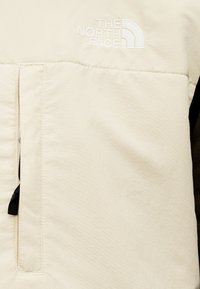 Ύφασμα σε κρεμ απόχρωση με κεντημένο λογότυπο The North Face και κάθετη τσέπη με φερμουάρ και μαύρο έλκτη.