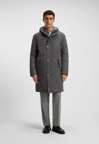 Grahamelierter Wollmischmantel mit Kapuze, Druckknopfverschluss und Seitentaschen, kombiniert mit hellgrauen Hosen und schwarzen Schuhen, zeigt eine strukturierte Silhouette.
