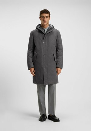 Cappotto in misto lana grigio con cappuccio, chiusura a scatto e tasche laterali, abbinato a pantaloni grigi chiari e scarpe nere, che mostrano una silhouette ben definita.