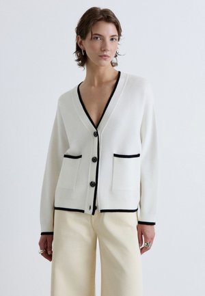 Donna che indossa un cardigan bianco con bottoni, rifinito in nero e con tasche, abbinato a pantaloni ampi color crema su uno sfondo semplice.