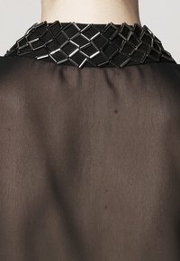 Top in tessuto trasparente nero con colletto metallico testurizzato e geometrico. Il colletto presenta link rettangolari argentati con un motivo a griglia.