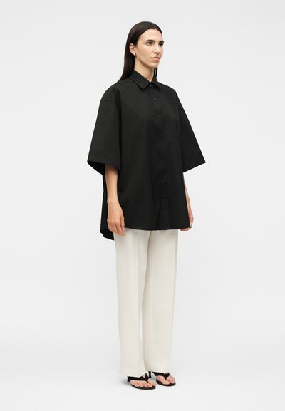 Γυναίκα που φοράει μαύρο φαρδύ πουκάμισο με κοντά μανίκια και κουμπιά, λευκό φαρδύ παντελόνι και μαύρα σανδάλια με ανοιχτά δάχτυλα, μπροστά σε απλό φόντο.