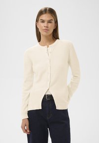 Donna che indossa un cardigan crema con bottoni sopra un top bianco, jeans scuri e una cintura nera, in piedi con una mano nella tasca.