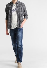 Svart och vit rutig skjorta över en ljusgrå t-shirt med tryck, parat med blå jeans och beige skor. Avslappnad outfit med ledig passform.