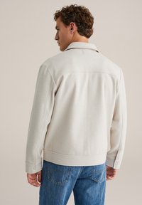 Chaqueta beige claro con cuello con solapa y puños con botones, fabricada con una tela suave de textura lisa y detalles mínimos.