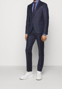 Marineblauw pak met twee knopen, notch revers en een slim fit. Gepaard met een lichtblauw overhemd en een donkere stropdas, gedragen met witte sneakers.