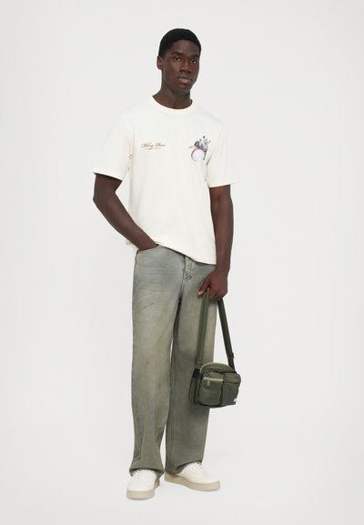 Man standing wearing off-white graphic t-shirt, faded wide-leg jeans, white sneakers, holding green crossbody bag by its straps.