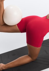 Personne en shorts rouges équilibrant un ballon d'exercice blanc texturé entre un genou plié et un bras sur un tapis d'exercice noir.