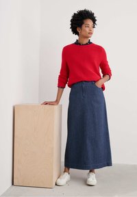 Un pull rouge côtel�é associé à une longue jupe en denim. La jupe présente une couture frontale, et elle porte des baskets blanches tout en s'appuyant sur un cube en bois clair.