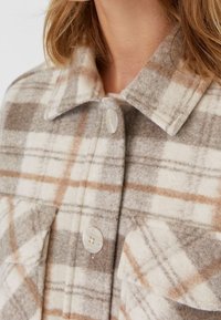 Chemise en flanelle à carreaux beige et gris mettant en avant un col, deux poches poitrine et des fermetures à boutons. Texture douce, avec des accents de boutons ronds.