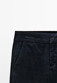 Sezione di pantaloni in corduroy neri con texture a coste, dotata di una cintura e tasche laterali. Il tessuto è morbido, con sottili linee verticali.