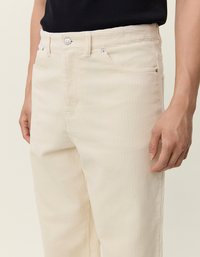 Beige kordbársony nadrág egyenes szabással, függőleges bordás textúrával, elülső zsebekkel és fémből készült gombos záródással.