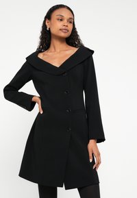 Sieviete ar cirtainiem matiem, tērpusies melnā, pogās aizvērtā mētelī ar platām apkaklēm, stāv ar vienu roku uz gurna uz baltas, vienkāršas fona.