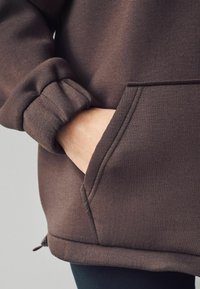 Main d'une personne dans la poche avant d'un sweat à capuche marron avec poignets élastiques et détails de couture visibles.