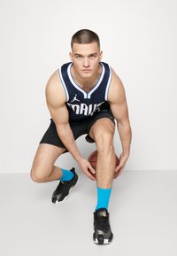 Basketballspiller i en marineblå trøje med hvidt trim og shorts, iført sorte sko og lyseblå sokker, knælende med en basketball i hånden.