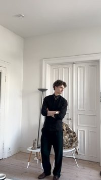 Svart skjorta med knappar, mörka vida byxor, bruna skor; modern inredning med vita väggar, trägolv och en dekorativ lampa och stol.