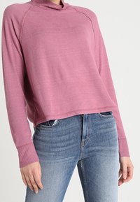 Rosa Pullover mit hohem Kragen, gerippten Bündchen und legerem Schnitt, kombiniert mit blauen Jeans im klassischen Fünf-Taschen-Design.
