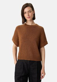 Brauner gestrickter Pullover mit kurzen Ärmeln, einem gerippten Saum und strukturierten Schultern, kombiniert mit schwarzen Hosen.