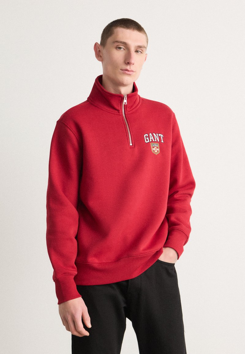 Hanorac roșu cu fermoar pe jumătate, confecționat din material fleece, având un logo pe piept, cu manșete elastice și tiv, asortat cu pantaloni negri.