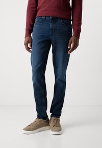 Dark blue denim jeans with a straight cut, worn with a maroon sweater and beige suede sneakers, displayed against a neutral background.