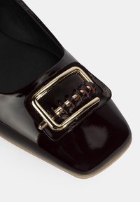 Chaussure en cuir verni bordeaux avec un embout pointu et un détail de boucle en métal doré, ornée d'accents tissés sur le dessus. Texture lisse et fini brillant.