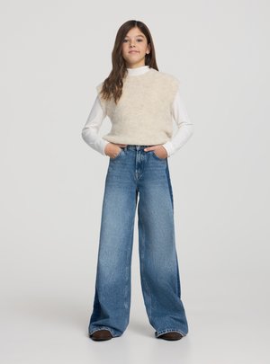 Ragazza con lunghi capelli castani indossa un maglione beige senza maniche sopra una maglia bianca a maniche lunghe e jeans blu a gamba larga con le mani nelle tasche.