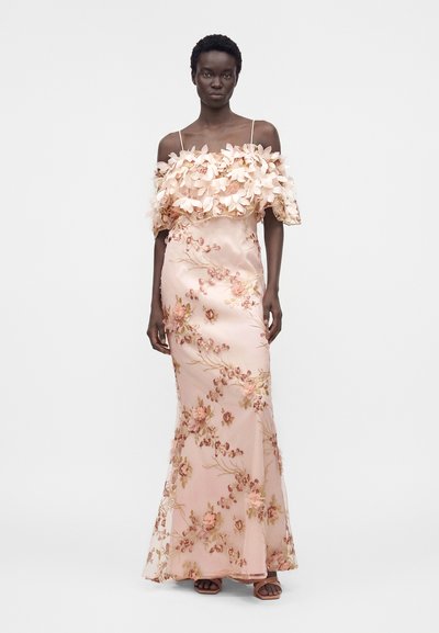 Γυναίκα με μακρύ φόρεμα σε ροζ απόχρωση με ρετρό ντεκολτέ και κεντητά λουλούδια, στέκεται μπροστά σε απλό φωτεινό φόντο.