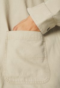 Beige canvas jacket with a visible hand in the patch pocket. The texture is slightly coarse, featuring exposed stitching and a cuff detail.