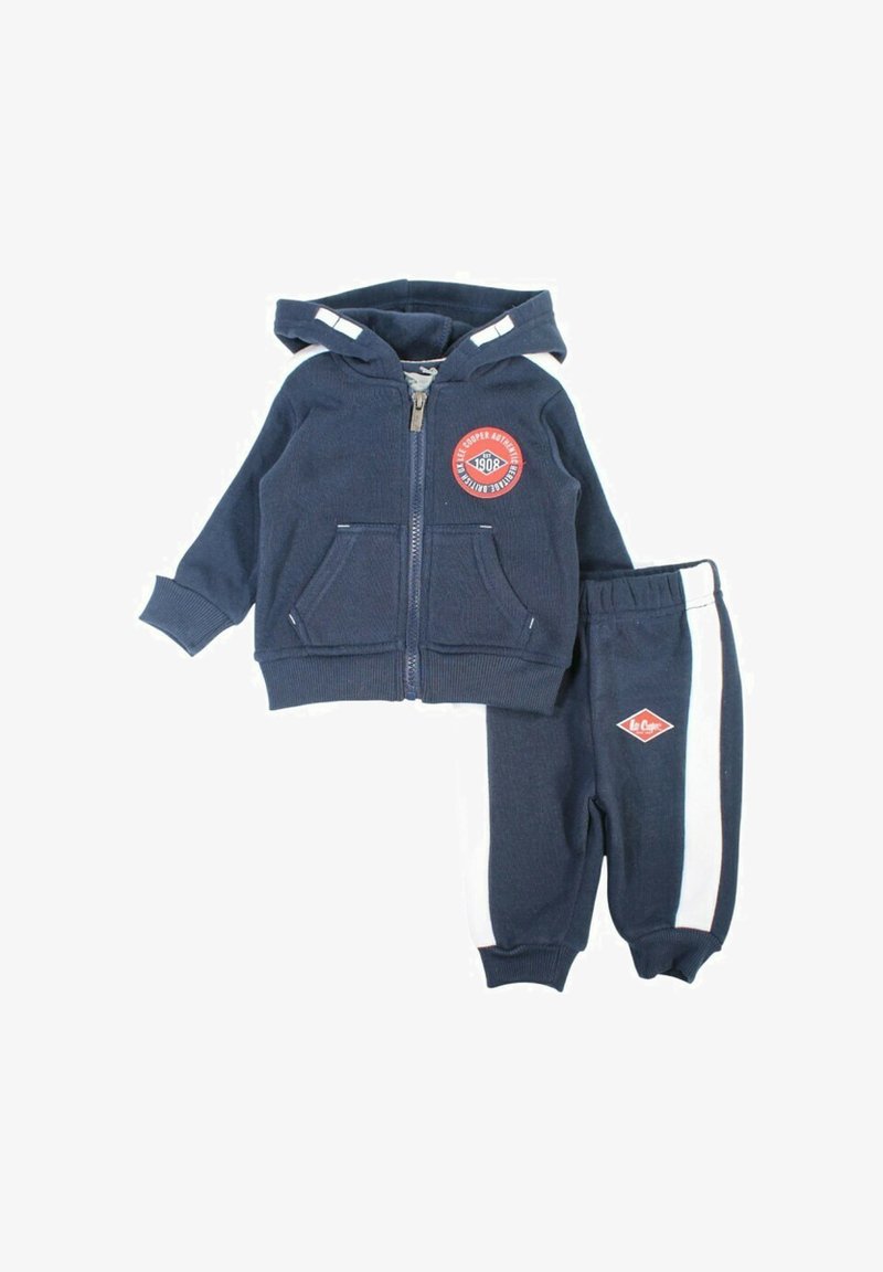 Survêtement pour tout-petit bleu marine comprenant un sweat à capuche zippé avec poches avant et un écusson logo, assorti à un pantalon avec des bandes blanches sur les côtés.